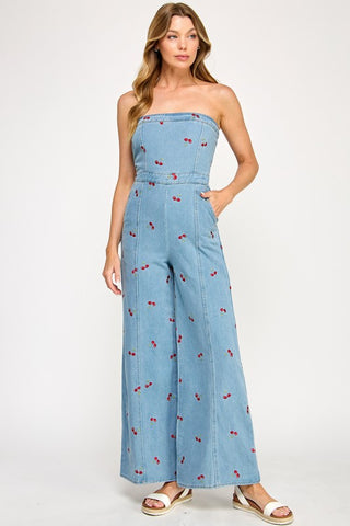 A woman wears The Vintage Shops Strapless Denim Cherry Jumpsuit, featuring embroidered cherries and wide-leg pants, paired with white sandals against a plain light background.