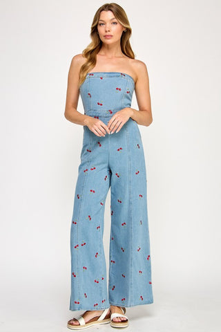 A woman wears The Vintage Shops Strapless Denim Cherry Jumpsuit, featuring a retro-inspired design with embroidered cherries and wide-leg pants, paired with white sandals against a plain white background.