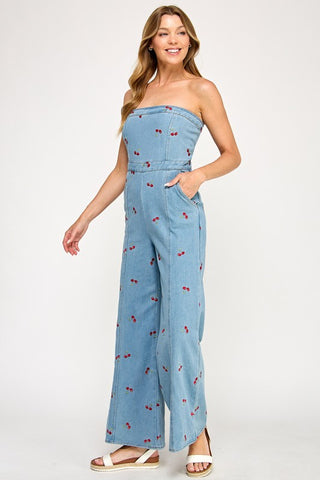 A woman smiles in The Vintage Shops Strapless Denim Cherry Jumpsuit featuring a retro cherry pattern and wide legs. She wears white sandals, has long wavy hair, and poses against a plain white background.