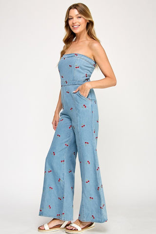 A woman smiles in The Vintage Shops Strapless Denim Cherry Jumpsuit, a retro-inspired, light blue piece with wide legs, embroidered cherries, and side pockets. She pairs it with white sandals and lets her wavy hair flow against a plain background.