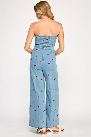 A woman faces away in The Vintage Shops Strapless Denim Cherry Jumpsuit featuring a retro cherry print, light blue hue, wide-leg cut, and strapless design. She has long blonde hair, wears white sandals, and stands against a plain white background.