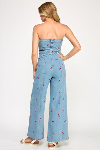 A woman with long blonde hair stands against a plain background wearing The Vintage Shops Strapless Denim Cherry Jumpsuit, a retro-inspired light blue piece featuring small embroidered cherries, styled with flat sandals.
