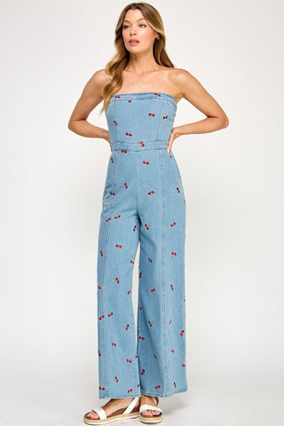 A woman with loose, wavy hair wears The Vintage Shops Strapless Denim Cherry Jumpsuit—featuring wide-leg pants and an embroidered cherry print—paired with white sandals against a plain background.