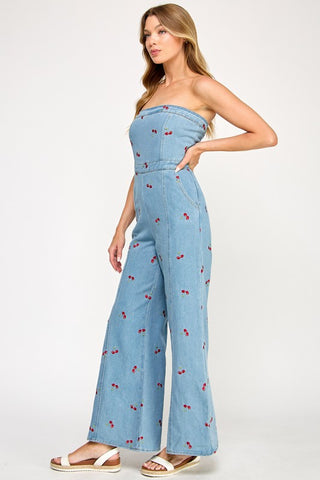A woman poses sideways with one hand on her hip, wearing The Vintage Shops Strapless Denim Cherry Jumpsuit and white sandals. Her long hair is loose, and she stands against a plain background.