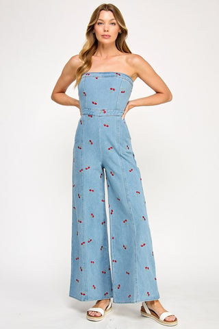 A woman poses against a white background in The Vintage Shop’s Strapless Denim Cherry Jumpsuit, featuring a retro-inspired cherry print, wide legs, and styled with white sandals. She has long, wavy hair.