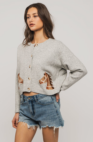 A woman stands against a plain background wearing the Urban Daisy Horse Graphic Cropped Cardigan, a light gray button-up sweater with horse motifs and ribbed trim, styled with denim cutoff shorts and one hand on her hip.