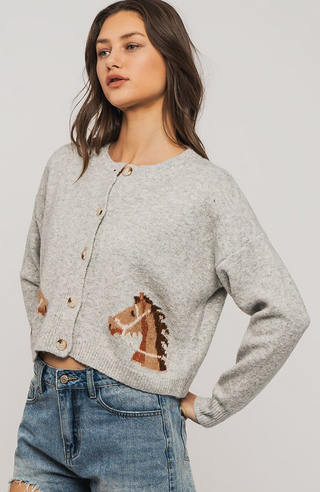 A woman models Urban Daisys Horse Graphic Cropped Cardigan, featuring brown horse head motifs and ribbed trim, styled with light blue ripped jeans against a plain background.