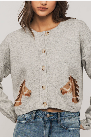 A woman wears the Urban Daisy Horse Graphic Cropped Cardigan, a light gray piece with brown and white horse motifs on each pocket and ribbed trim, paired with blue jeans. Her face is partially out of frame.