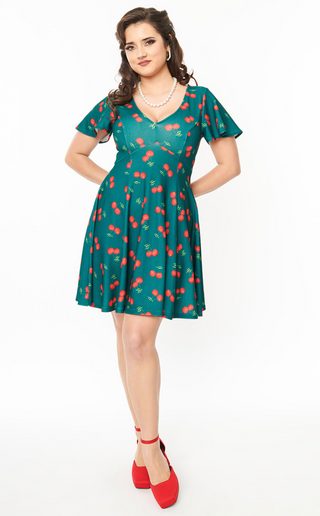 A woman wears the Unique Vintage Fit & Flare Cherry Dress with red shoes and a pearl necklace, smiling with her hands behind her back against a plain white background.