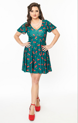A woman in the Unique Vintage Fit & Flare Cherry Dress, paired with a pearl necklace and red heels, stands confidently with hands on hips against a plain white background.