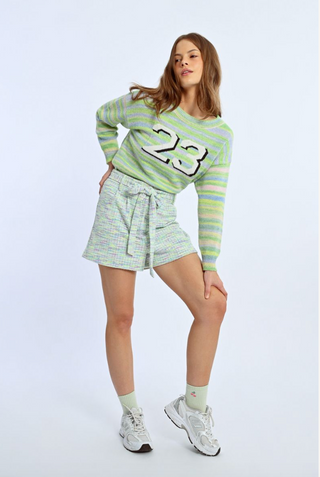 A woman models Molly Brackens Knit Self-Tie Belt Shorts with a light green and lavender striped number 23 sweater, light socks, and white sneakers against a plain light background.