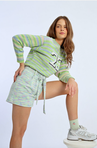 A woman poses confidently with one leg raised on a stool, wearing Molly Bracken Knit Self-Tie Belt Shorts, a striped green and purple sweater with the number 25, light green socks, and gray sneakers against a plain background.