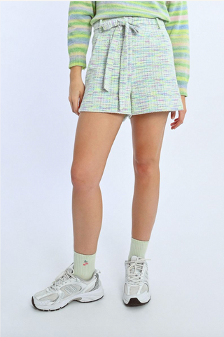 A person in Molly Bracken Knit Self-Tie Belt Shorts, a green and pastel-striped sweater, pale green socks, and white sneakers stands against a plain light background.