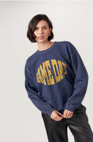 A woman with short dark hair wears the Pros Game Day Vintage Sweatshirt in navy peony blue with bold yellow GAMEDAY lettering and black leather pants, standing against a plain light background.