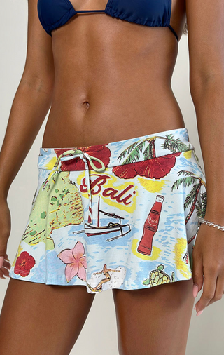 A person wears a navy bikini top and the Motel Rocks Saeko Micro Skort, featuring a Bali-inspired print with palm trees, flowers, sailboat, Coca-Cola bottle, and bold red Bali lettering.
