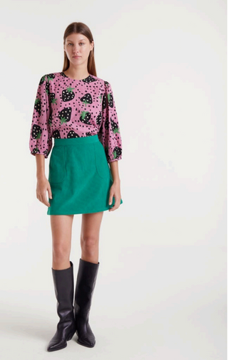 A woman stands against a plain background wearing a pink blouse with black polka dots and hearts, the Compania Fantastica Corduroy Mini Skirt in green, and knee-high black boots.