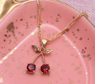 The Sweet Crystal Cherry Gold Necklace by Zad features a gold chain with a cherry pendant, two red rhinestones, and delicate leaf accents, elegantly displayed on a pink dish with gold speckles.