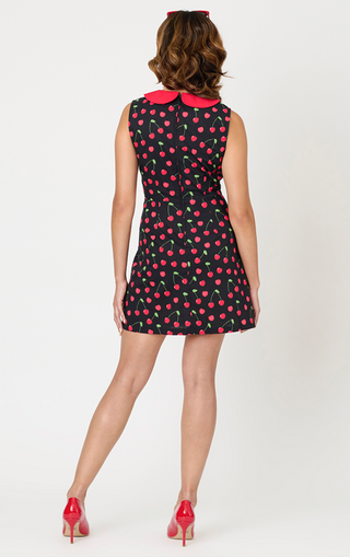 A woman with wavy brown hair faces away in a Smak Parlour Sleeveless Cherry Print Shift Dress, featuring a striking red collar. She wears red heels. Available in sizes XS-XL. The background is plain white.