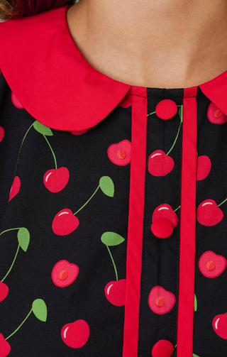 Close-up of the Smak Parlour Sleeveless Cherry Print Shift Dress (sizes XS-XL), featuring a black base, red collar and trim, red buttons, and a playful cherry pattern with green stems.