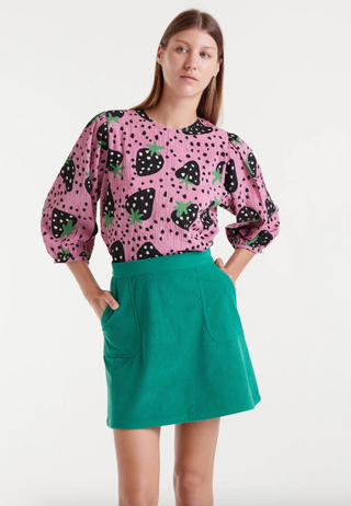 A woman stands with her hands in the pockets of a green skirt, wearing the Compania Fantastica Morango Strawberry Print Top, featuring a pink long sleeve design with black and green strawberry print, against a plain white background.