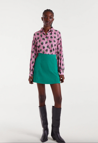 A woman stands against a white background wearing a pink patterned long-sleeve blouse, black knee-high boots, and the Compania Fantastica Corduroy Mini Skirt in green with front pockets.