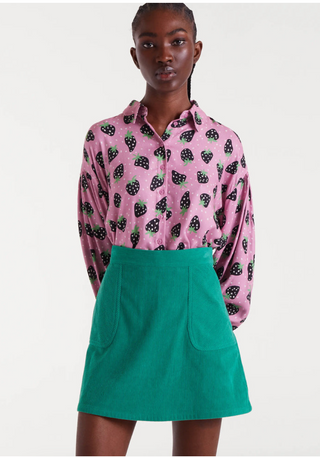 A woman poses against a plain background wearing a pink button-up shirt with a black and white fruit pattern, tucked into the Compania Fantastica Corduroy Mini Skirt—a high-waisted green skirt with front pockets.