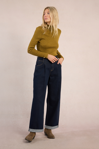 A woman stands against a plain beige background, wearing the Molly Bracken High Neck Knit Sweater in mustard yellow and high-waisted wide-leg dark blue jeans with cuffs, paired with brown suede ankle boots. She looks left, hands in pockets.