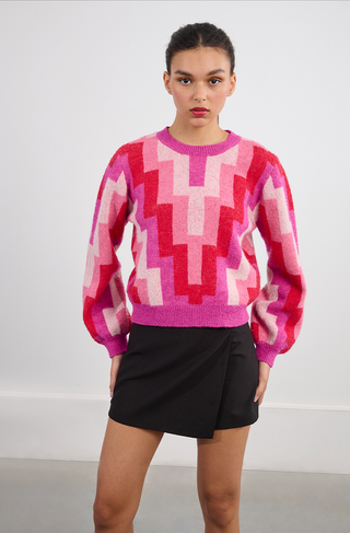 A young woman wears the Molly Bracken Retro 80s Knit Sweater, featuring a colorful geometric pattern in pink and red, paired with a black wrap mini skirt. Her hair is pulled back as she looks directly at the camera with a neutral expression.