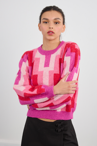 A woman with dark hair tied back wears the Molly Bracken Retro 80s Knit Sweater in pink, red, and purple geometric patterns. She stands against a plain white background with arms crossed and a neutral expression.
