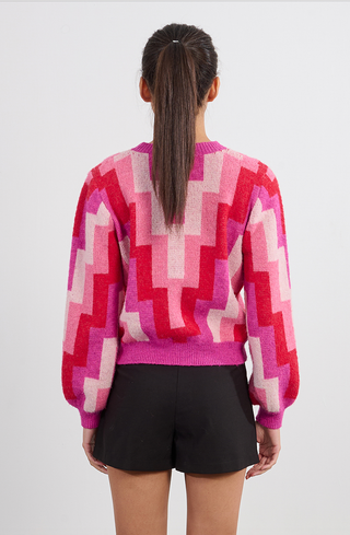 A woman with a ponytail, seen from behind, wears the Molly Bracken Retro 80s Knit Sweater—featuring a colorful geometric pink, red, and white pattern—with black shorts against a plain light background.