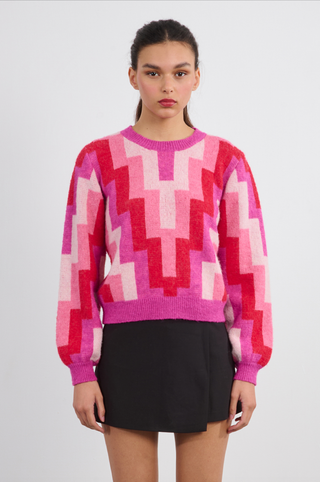 A young woman with tied-back hair and a neutral expression models the Molly Bracken Retro 80s Knit Sweater—featuring vibrant pink and red geometric patterns—paired with a short black skirt against a plain background.