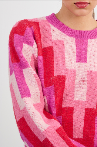 A person is wearing the Molly Bracken Retro 80s Knit Sweater, featuring a pink and red geometric pattern; only their lower face and upper torso are visible.