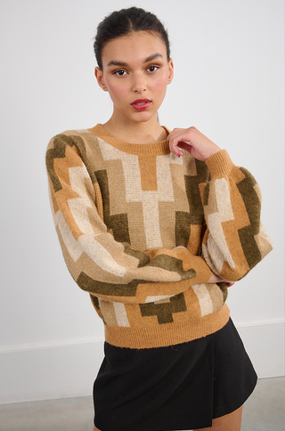 A young woman poses indoors against a white wall, wearing the Molly Bracken Retro 80s Knit Sweater featuring geometric patterns in brown, beige, and cream, paired with a black skirt. She touches her chin thoughtfully.