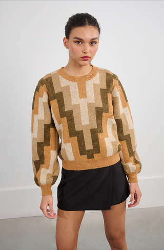 Indoors, a person wears the Molly Bracken Retro 80s Knit Sweater featuring brown, beige, and olive green zigzag patterns with a short black skirt. Their hair is tied back as they look slightly past the camera.