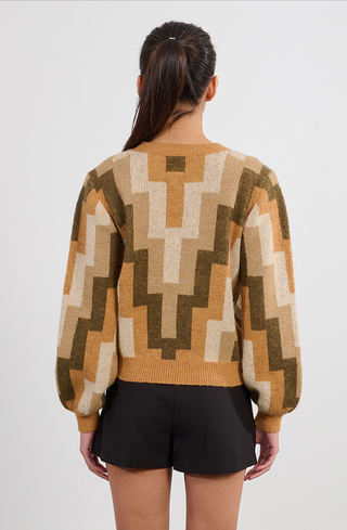 A woman with long dark hair in a ponytail wears the Molly Bracken Retro 80s Knit Sweater in beige, tan, and green tones with black shorts, standing facing away from the camera against a plain background.
