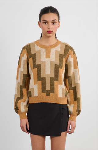 A young woman with dark hair in a ponytail wears the Molly Bracken Retro 80s Knit Sweater in tan, beige, and olive tones, paired with a black mini skirt. She stands against a plain white background looking forward neutrally.