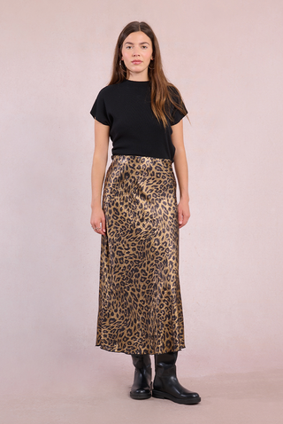 A woman with long brown hair and a neutral expression wears a black short-sleeve top, black ankle boots, and the Molly Bracken Gold Metallic Leopard Midi Skirt, standing against a light pink background.