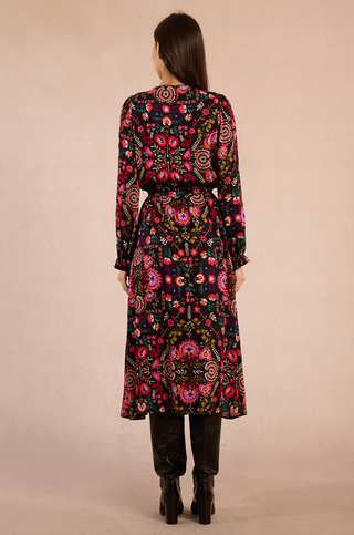 A woman with long brown hair faces away, wearing the Molly Bracken Irina Print Maxi Dress with a vibrant floral pattern and black knee-high boots, set against a plain beige background.