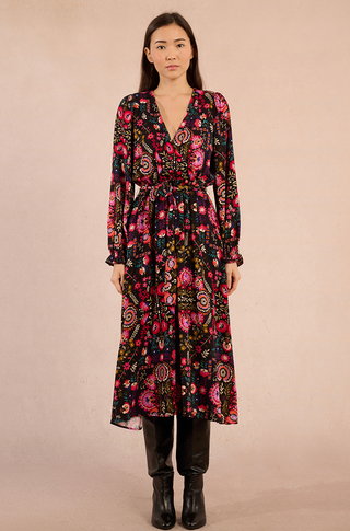 A woman stands against a beige backdrop, wearing Molly Brackens Irina Print Maxi Dress—an ankle-length, long-sleeved black floral piece in soft viscose with a cinched waist—paired with black knee-high boots.