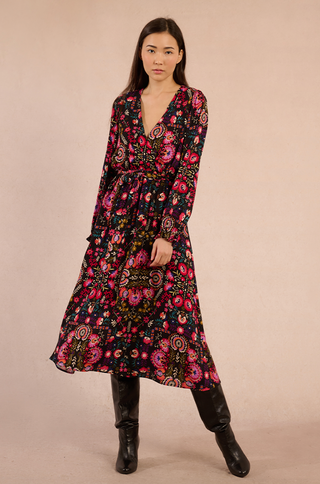 Irina wears the Molly Bracken Irina Print Maxi Dress, an ankle-length black multicolor viscose dress with bright pink and red florals, paired with black knee-high boots. She stands against a beige background with straight brown hair and a neutral expression.