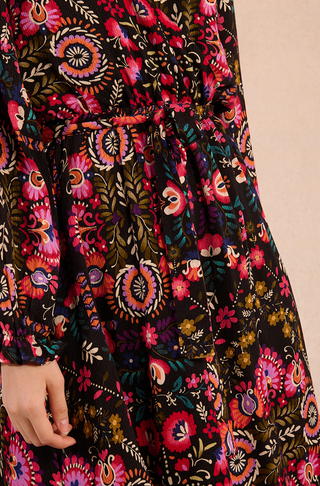 Close-up of a person wearing the Molly Bracken Irina Print Maxi Dress, featuring colorful floral and paisley patterns on soft viscose, a tied waist belt, and long sleeves, with only part of the arm and hand in view.