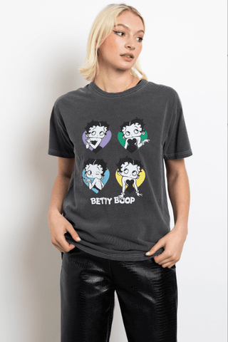 A woman with blond hair models the Daisy Street Betty Boop Hearts Graphic Tee-FINAL SALE in charcoal gray, featuring four colorful Betty Boop illustrations, styled with shiny black pants against a white background.