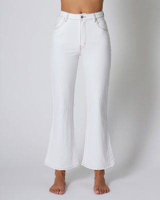 A person models Rollas Classic Straight Ankle Denim in 80s White with bare feet against a plain light gray background. The high-waisted jeans feature front pockets, a button closure, and a zip fly.
