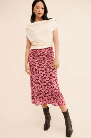A woman with long dark hair and a neutral expression stands against a beige background wearing a white sleeveless top, the MINKPINK Dakota Mesh Midi Skirt-FINAL SALE with a ruched waist, and black ankle boots.