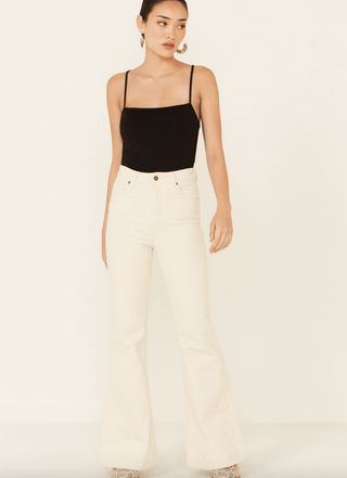 A woman stands against a plain background in Rollas East Coast Flare Corduroy-FINAL SALE jeans and a black spaghetti-strap top, her hair tied back with earrings, showcasing an effortless coastal style.