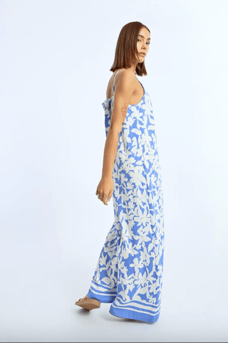 A woman with straight brown hair models the Molly Bracken Floral Strappy Jumpsuit-FINAL SALE, a long blue floral print piece with white-and-blue striped hem, standing sideways against a plain light background.