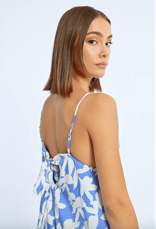 A woman with straight, shoulder-length brown hair models the Molly Bracken Floral Strappy Jumpsuit-FINAL SALE in blue with white floral patterns, thin straps, and a back bow. She glances over her shoulder against a light background.