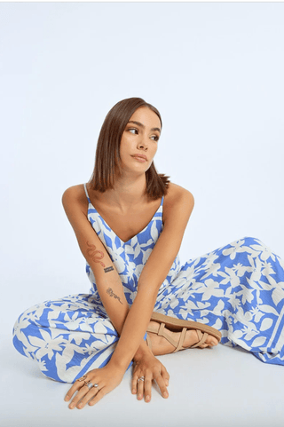A woman with straight brown hair and tattoos sits on the floor in a Molly Bracken Floral Strappy Jumpsuit-FINAL SALE, paired with tan sandals and rings. She looks to the side against a plain light blue background.