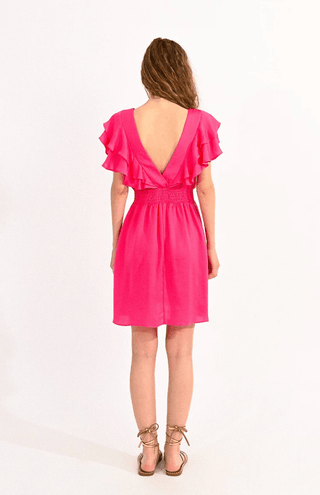 A woman with wavy brown hair and tan sandals faces away, modeling the Molly Bracken Flutter Ruffle Mini Dress-FINAL SALE in hot pink, featuring a deep V-back, ruffled short sleeves, and cinched waist against a white background.