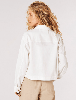 A woman with wavy blonde hair faces away, wearing the Apricot Lightweight Crop Jacket-FINAL SALE in white cotton blend and beige pants, set against a plain light background.